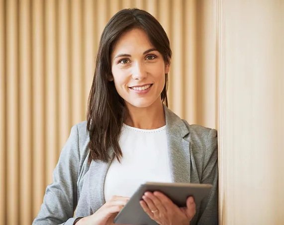 A smiling woman in a business suit holding a tablet in front of a wooden wall.
